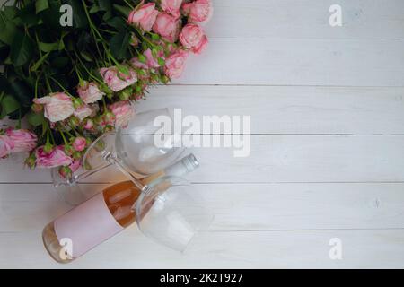 Un grande bouquet di rose rosa e una bottiglia di vino rosato con bicchieri giacciono sul lato su uno sfondo bianco di legno con un posto per il testo Foto Stock