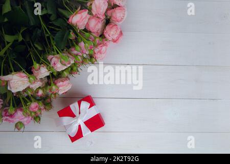 Un grande bouquet di rose rosa che fioriscono e gemme si trova e un regalo in una casella rossa sul lato su uno sfondo bianco di legno con un posto per il testo Foto Stock