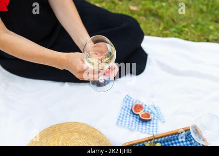 Romantico pic-nic nel parco sull'erba, cibo delizioso: Cesto, vino, uva, fichi, formaggio, tovaglia a scacchi blu, due bicchieri di vino. La ragazza sta tenendo un bicchiere di vino nelle sue mani.il concetto di ricreazione all'aperto. Foto Stock