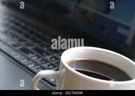 Una tazza bianca con caffè nero primo piano sullo sfondo di un notebook nero. Concetto aziendale Foto Stock