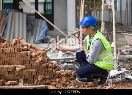 Appaltatore asiatico con casco di sicurezza, giubbotto riflettente e stivali di gomma che controlla la qualità del mattone mentre tiene walkie talkie. Giovane ingegnere che lavora in cantiere. Foto Stock