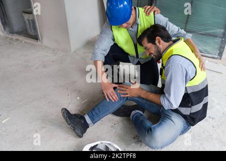 Il giovane uomo caucasico in giubbotto di sicurezza gli afferrò il ginocchio e urla nel dolore. A causa di lesioni in cantiere. Il suo amico asiatico viene e lo aiuta a stare in piedi. Foto Stock