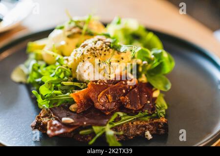 Uova benedict con prosciutto secco e insalata mescolate su un piatto nero. Vista ravvicinata, messa a fuoco selettiva. Foto Stock