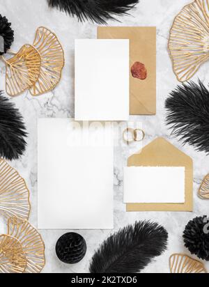 Cartoncini e busta su tavola in marmo vicino a piume nere e foglie dorate. Mockup matrimonio Foto Stock