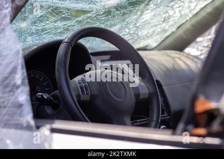 Il volante di una vettura dopo un incidente. Gli airbag lato conducente non si sono attivati. Vista dalla finestra laterale sinistra. Parabrezza rotto con volante. Cruscotto e volante neri polverosi. Foto Stock