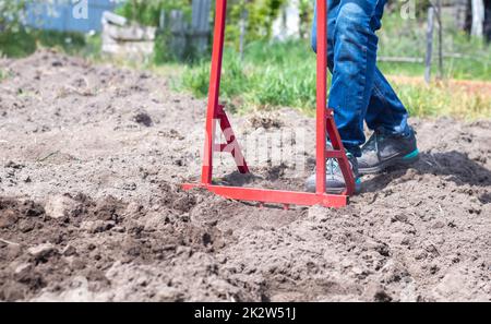 Un contadino in jeans scava il terreno con una pala a forcella rossa. Una pala miracolosa, uno strumento pratico. Coltivatore manuale. Il coltivatore è un efficiente strumento manuale per il dissodamento. Allentamento del letto. Foto Stock