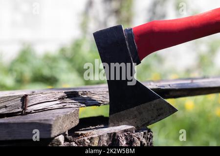 Una nuova ascia moderna con manico rosso si stende da un ceppo di legno contro un prato verde in una giornata di sole d'estate. L'asse si è incastrato nel moncone. Lama AX in un log. Foto Stock