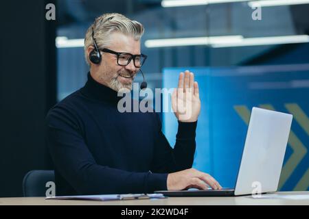 Incontro online con colleghi, uomo senior dai capelli grigi che lavora in un moderno ufficio interno, dirigente con cuffie e occhiali sorridenti e guardando la fotocamera del computer portatile che sventolano il gesto di saluto, videochiamata Foto Stock