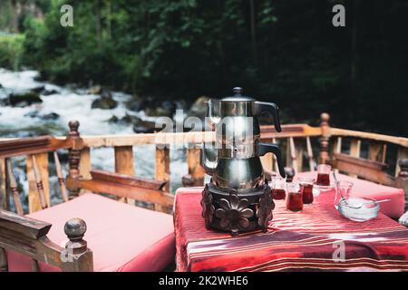 Vista frontale del tradizionale tè turco cromo bollitore e bicchieri serviti su un tavolo da ristorante con sedie vicino a un fiume in un ambiente esterno Foto Stock