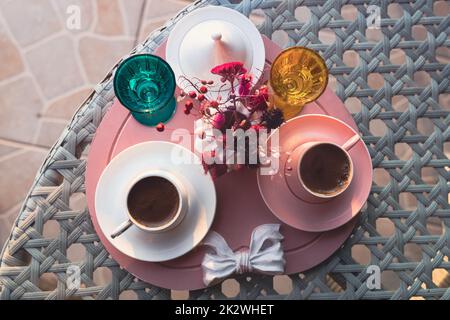 Primo piano con vista frontale delle tradizionali tazze di caffè turche servite all'esterno con fiori d'acqua e dessert deliziati in una romantica preparazione su un tavolo di vetro al tramonto Foto Stock