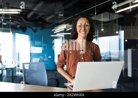 Ritratto di giovane bella donna ispanica con notebook, donna d'affari all'interno di un moderno edificio ufficio sorridendo e guardando la macchina fotografica. Foto Stock