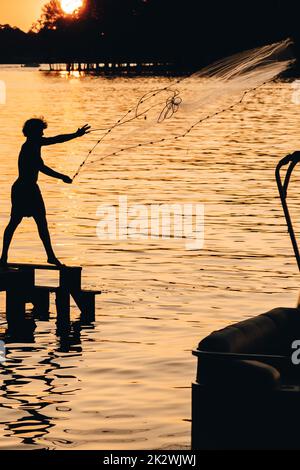 Uno scatto verticale di una silhouette di una persona che pesca con una rete in un lago al tramonto Foto Stock