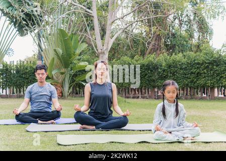 madre, padre che pratica le esercitazioni di yoga con la figlia del bambino all'aperto nella posa meditate insieme Foto Stock