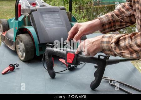 Un uomo sta riparando un rasaerba alimentato a batteria. L'interruttore a levetta elettrico per avviare il motore è difettoso e deve essere sostituito. Un artigiano al lavoro. Foto Stock