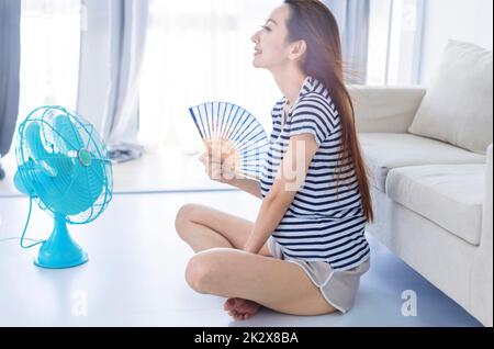 La giovane donna si rilassa allagata in soggiorno con ventilatore a mano e godendo del flusso d'aria dal ventilatore . Concetto di calore estivo Foto Stock