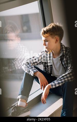 Triste adolescente in abiti casual guarda fuori dalla finestra mentre si siede sul pavimento all'interno Foto Stock