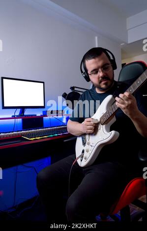 produttore di musica rock maschile bearded in occhiali e cuffie, registrando la chitarra elettrica seduta nel suo studio di musica di casa. Foto Stock