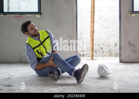 Il giovane uomo caucasico in gilet di sicurezza gli afferrò il ginocchio e urla nel dolore. A causa di lesioni nel cantiere. Foto Stock