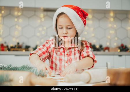 Piccola ragazza dai capelli scuri di 3 anni con berretto rosso di Natale e camicia a scacchi taglia i biscotti di pan di zenzero dall'impasto arrotolato nel Natale bianco Foto Stock