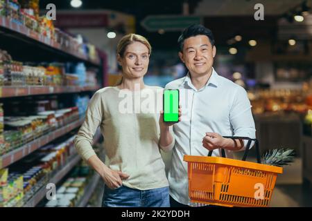 Giovane famiglia diverse coppie di acquirenti nel supermercato, sorridendo e guardando la macchina fotografica, il reparto di alimentari, uomo e donna tenendo il carrello della spesa e mostrando schermo verde dello smartphone. Foto Stock