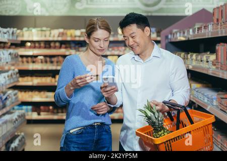 Famiglia diversa coppia asiatica uomo e donna in supermercato soddisfatto e felice shopping, utilizzando app sul telefono per selezionare i prodotti e la lista della spesa, utilizzare la carta di credito. Foto Stock