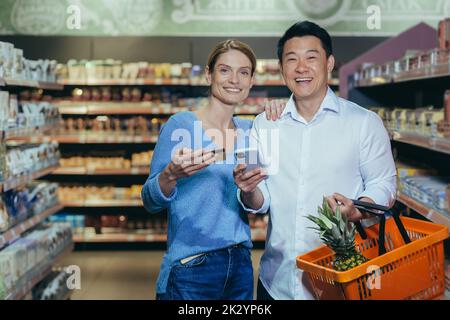 Felice coppia di famiglia diversa uomo e donna acquirenti in supermercato, guardando la macchina fotografica e sorridente shopping felice, tenendo in mano il telefono e la carta di credito bancario, con il carrello della spesa. Foto Stock