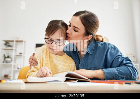 Ritratto di madre baciando figlia con sindrome di Down durante la lettura del libro insieme in una lezione di homeschooling Foto Stock