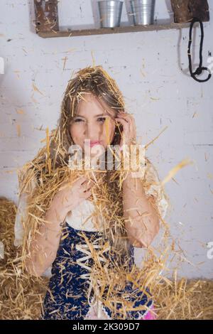 bella giovane donna, vestita in un abito contadino nel fienile giocando con il fieno gettandolo sul suo corpo, mestieri di fattoria Foto Stock