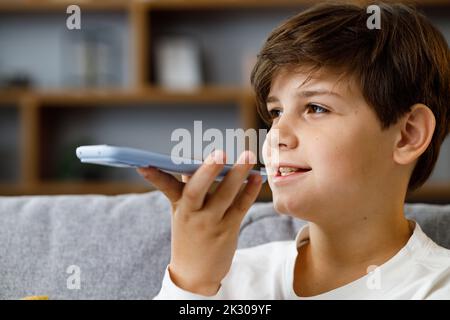 Giovane ragazzo carino che registra un messaggio vocale sullo smartphone. Sorridente adolescente che utilizza l'app Virtual Assistant seduto a casa in pullman. Riconoscimento vocale, audio Foto Stock