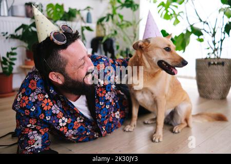 Uomo felice che indossa cappello da festa e occhiali da sole sdraiati con il cane a casa Foto Stock