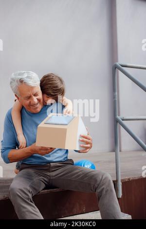 Uomo anziano sorridente che tiene modello di casa solare da nipote di fronte al muro Foto Stock