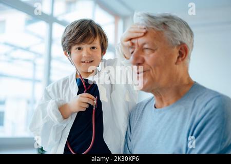 Ragazzo sorridente imitato come medico che controlla la temperatura del nonno a casa Foto Stock