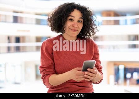Donna d'affari sorridente con capelli ricci che tiene lo smartphone in ufficio Foto Stock