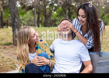 Donna felice che copre gli occhi dell'uomo seduto da un amico su panca al parco Foto Stock