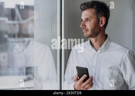 Uomo d'affari premuroso che tiene il telefono cellulare guardando attraverso la finestra Foto Stock
