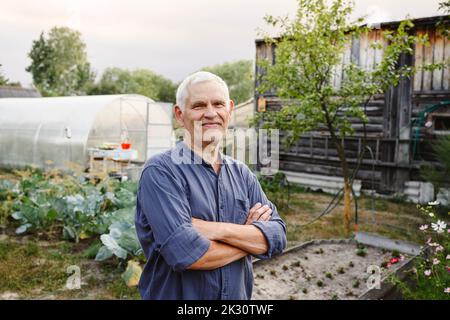 Uomo anziano sorridente in piedi con le braccia incrociate in giardino Foto Stock
