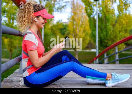 Sorridente sportswoman che tiene in mano il telefono cellulare per ascoltare la musica attraverso le cuffie auricolari wireless sulla passerella Foto Stock