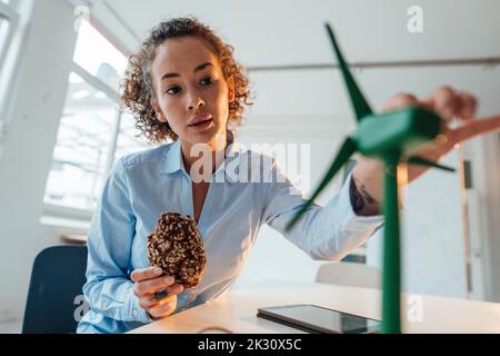 Un giovane ingegnere che tiene un biscotto che esamina il modello di turbina eolica alla scrivania in ufficio Foto Stock