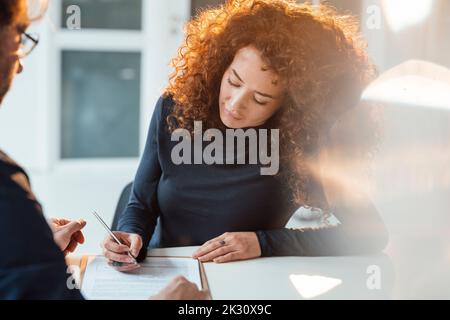 Donna d'affari con riccio capelli firma documento seduto alla scrivania in ufficio Foto Stock