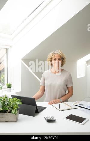 Donna d'affari con capelli ricci in piedi presso la scrivania in ufficio a casa Foto Stock