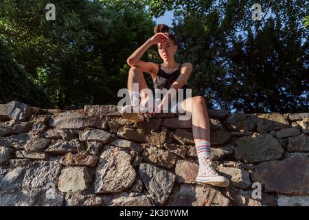 Hipster non binario seduto su un muro di pietra Foto Stock