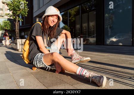 Sorridente persona non binaria seduta sullo skateboard sul sentiero Foto Stock