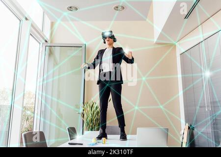 Donna d'affari che indossa un simulatore di realtà virtuale in piedi sul tavolo in ufficio Foto Stock