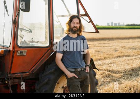 Sorridente contadino con le mani in tasca appoggiate al trattore in azienda Foto Stock