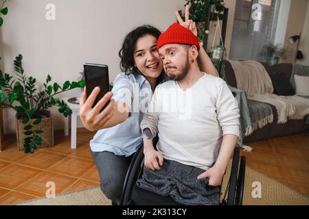 Donna felice con uomo in sedia a rotelle che prende selfie attraverso il telefono cellulare a casa Foto Stock