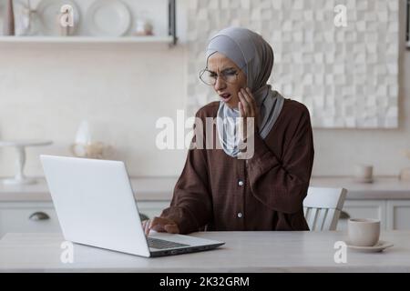 Donna musulmana mal di denti a casa, donna d'affari in hijab lavorare a distanza da casa, seduto in cucina con un computer portatile. Foto Stock