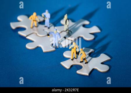 Squadra di minuscoli lavoratori in miniatura figure su jigsaw collegati puzzle pezzi isola su carta blu. Luce spettacolare con ombre lunghe. Foto Stock