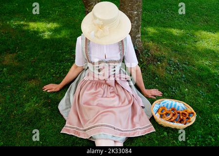 Una donna in un bel vestito tradizionale bavarese dirndl seduto sul prato verde con un cesto di pretzel o Brezeln (Monaco, Baviera) Foto Stock