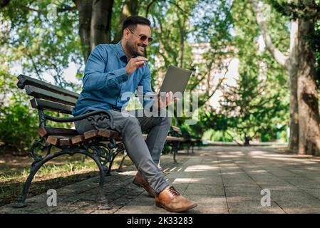 Immagine di un bell'uomo adulto, seduto su una panchina, che ha avuto un incontro con i suoi colleghi sul computer portatile. Foto Stock