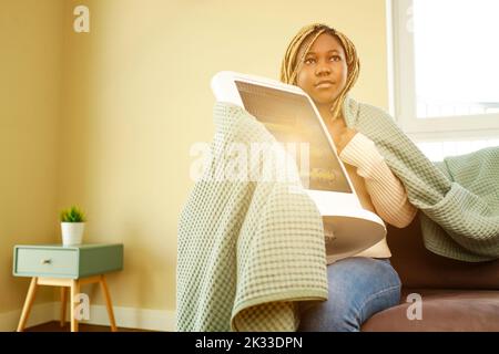 Donna afroamericana in coperta blu riscaldamento vicino al riscaldatore elettrico a casa Foto Stock